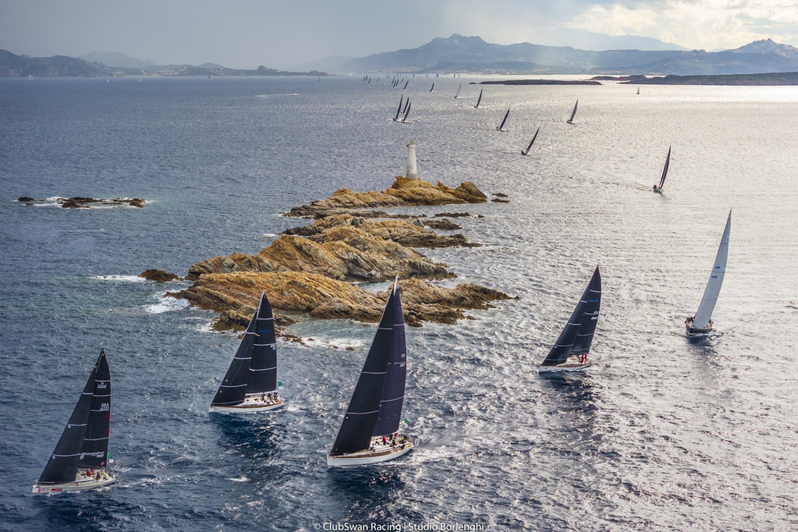 La flota de veleros Swan en regata, con la costa montañosa de Cerdeña al fondo. Foto: Studio Borlenghi / ClubSwan Racing